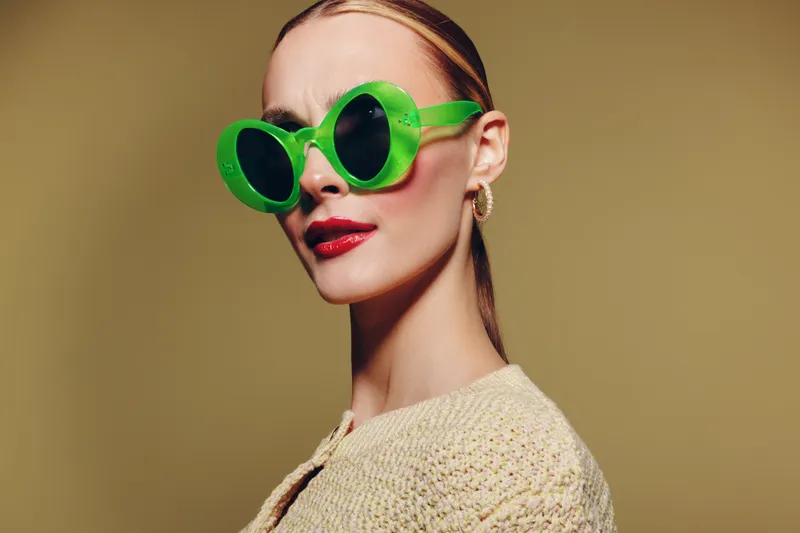 Stylish woman wearing large green sunglasses and red lipstick poses confidently against a beige background. Fashion concept with bold accessories and modern makeup style.