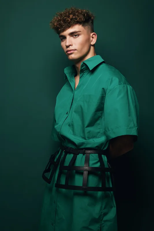 Young man with curly hair wearing green oversized shirt dress and unique black strap belt posing against green background, fashion model portrait with clean and modern style.