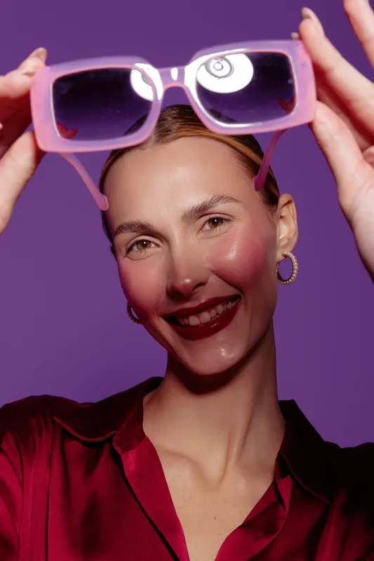 Smiling woman holds pink sunglasses in front of purple background wearing red blouse and gold hoop earrings. Concept of fashion accessories, summer style, and joyful mood.