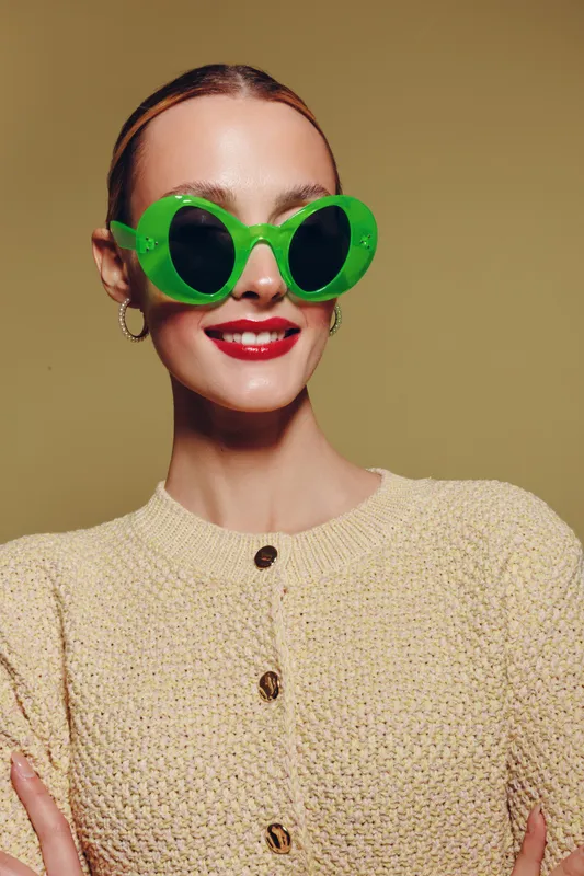 Smiling woman with bright red lipstick wears large green sunglasses and beige knitted sweater. Studio portrait on olive background. Concept of fashion, style, and confident attitude.