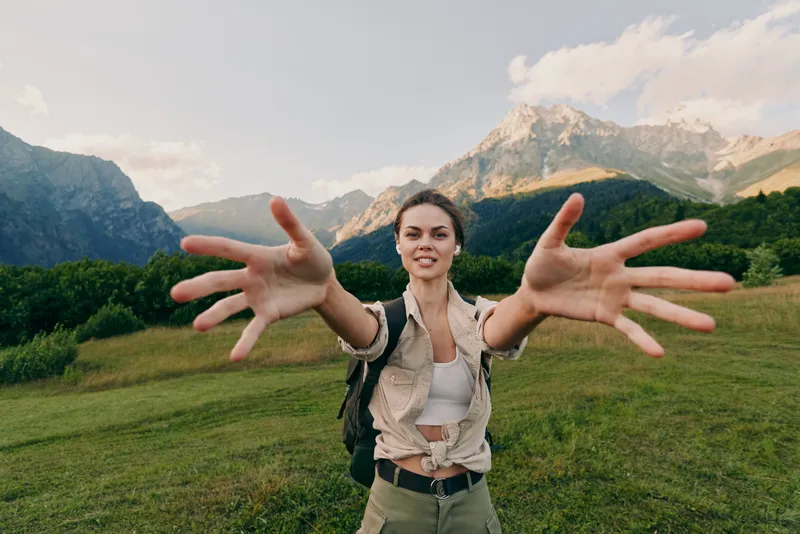Young woman with backpack extends arms toward the camera in a lush alpine meadow, mountains in the distance, conveying freedom and invitation to explore. Bright sky, relaxed smile.