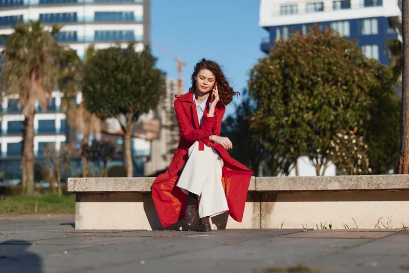 Young woman in a red coat sits on a stone bench outdoors talking on a smartphone with a calm expression on a sunny day in an urban park.