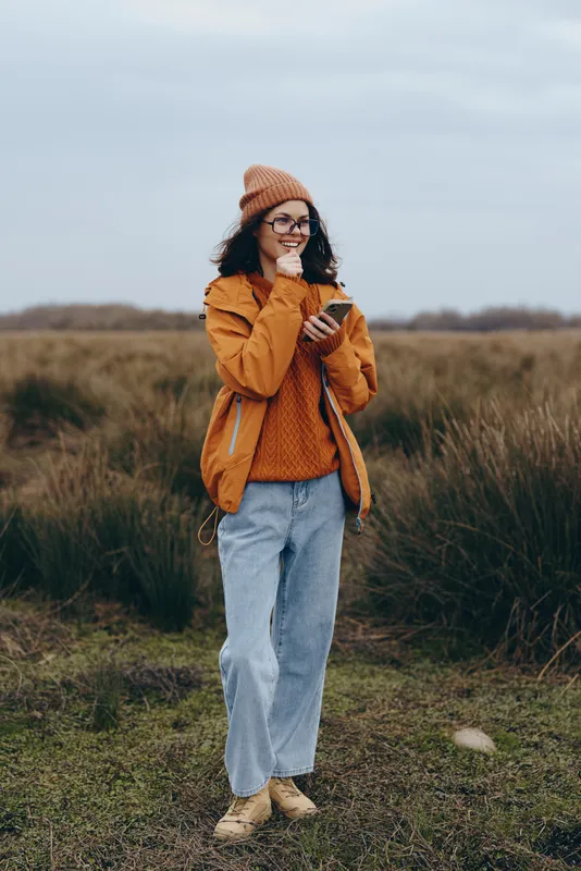 Woman outdoors in a grassy field, wearing orange jacket, knit beanie and jeans, smiling warmly, casual lifestyle, autumn light, cloudy sky, standing pose, natural colors