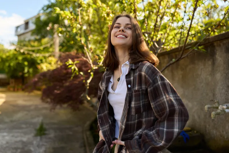 A young woman in plaid shirt smiles brightly outdoors in a sunlit garden, enjoying a carefree moment