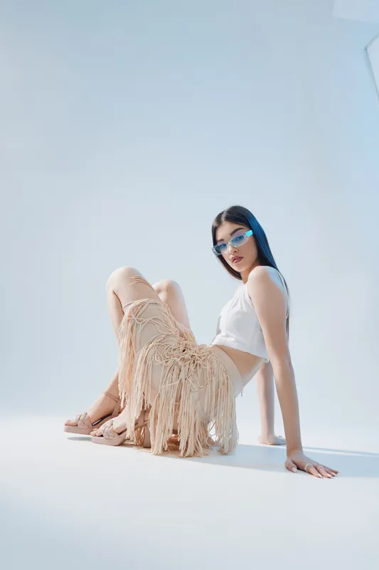Young woman in stylish fashion outfit poses sitting on the floor with blue sunglasses and fringed skirt. Minimalist studio with soft lighting highlights beauty and modern style.
