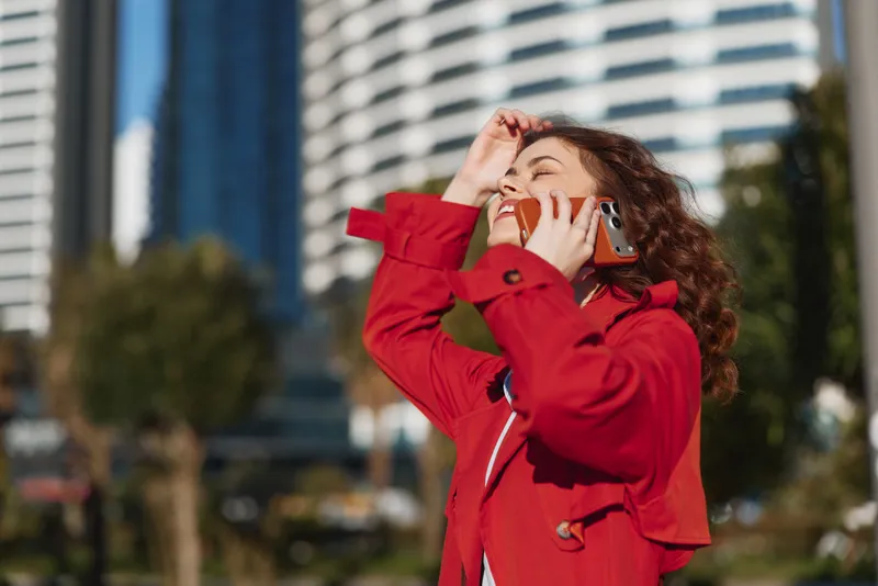 Red coated woman on a city street talks on a smartphone under bright daylight with tall glass buildings in the background
