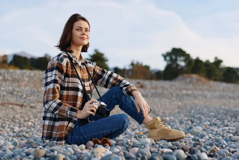 Woman photographer sitting on a rocky shore with a camera, wearing a plaid shirt and boots, a calm outdoor portrait among pebbles and distant trees