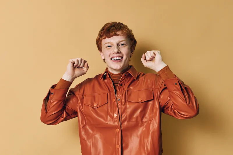 Young man with red hair smiling and stretching arms raised against plain beige background in casual shirt for positive energy and relaxation.