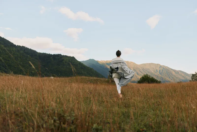 Woman walking through a tall grass field toward distant mountains, embracing outdoor living, calm sunlight, free spirit, natural landscape, serene mood, in the golden hour breeze quiet sky above.