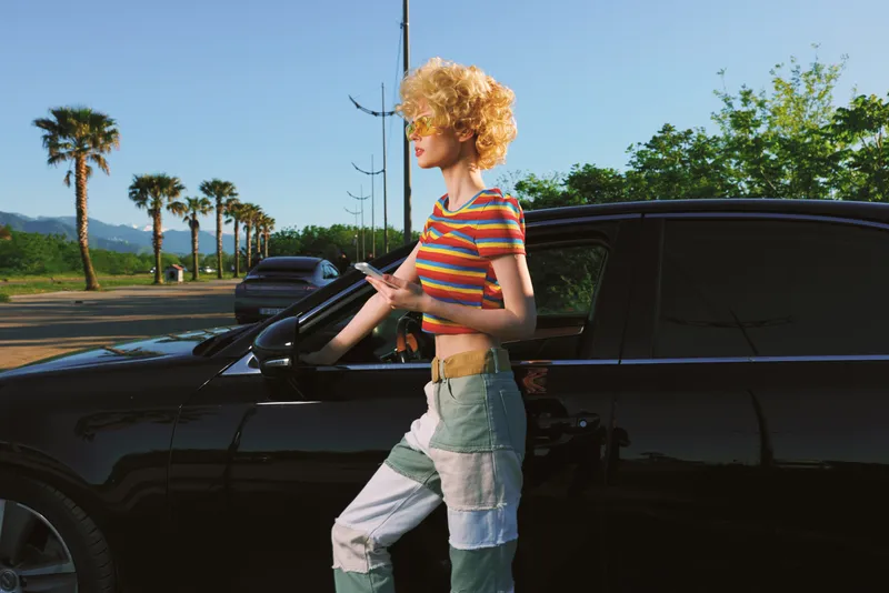 A stylish woman with curly hair leans on a sleek car in bright outdoor light, wearing a striped top and colorblock pants. A fashion moment capturing casual street style and bold color.
