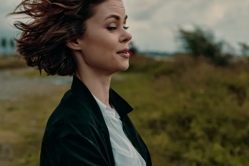 Profile of a woman outdoors with wind in hair, calm expression, wearing a dark jacket and light shirt against a blurred natural background, conveying freedom, movement, and confident mood in a candid