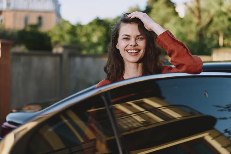 Smiling woman leaning from a car window outside, lifestyle, joyful expression, natural light, warm colors, candid portrait with bright reflections and soft shadows