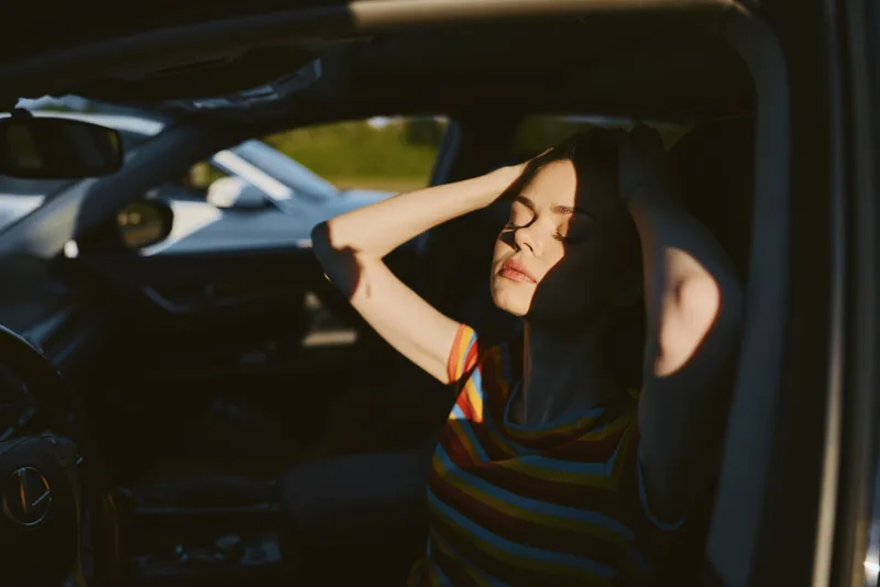 Smiling woman sitting inside a car, shielding eyes from bright sunlight, casual striped hoodie conveys relaxed lifestyle and sunny mood, retro film color, warm cinematic light enhances nostalgia and