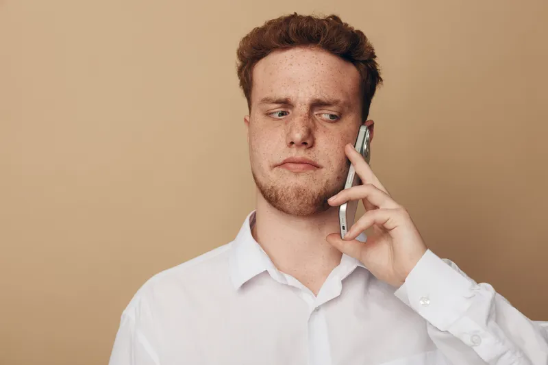 Young man in white shirt looks suspicious while talking on smartphone. Isolated on beige background. Concept of doubt, communication, and uncertain conversation.