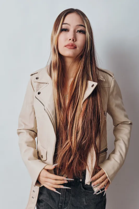 Young woman with long straight hair wearing beige leather jacket and black jeans standing against white background with confident serious expression and hands on waist.