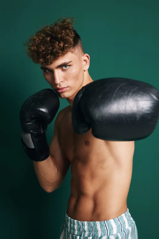 Young male boxer with curly hair wearing black gloves and striped shorts training in fighting stance against green background. Athletic, focused and muscular sportsman in action.