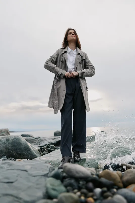 Young woman in long coat and wide trousers stands on rocky shore with waves splashing behind her. Overcast sky creates moody coastal atmosphere and fashion concept.