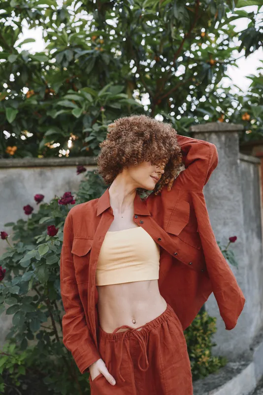 A woman with curly hair poses outdoors in a rust colored casual outfit, crop top with matching jacket and pants. Bright sunlight highlights textures and movement, conveying relaxed confidence