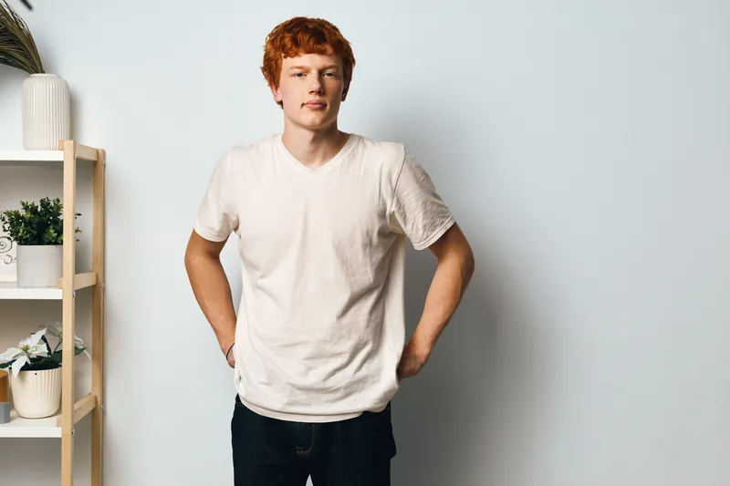 Man with red hair and casual white tshirt standing with hands behind back near wooden shelf with plants in minimalist room. Young male posing with neutral expression by wall.