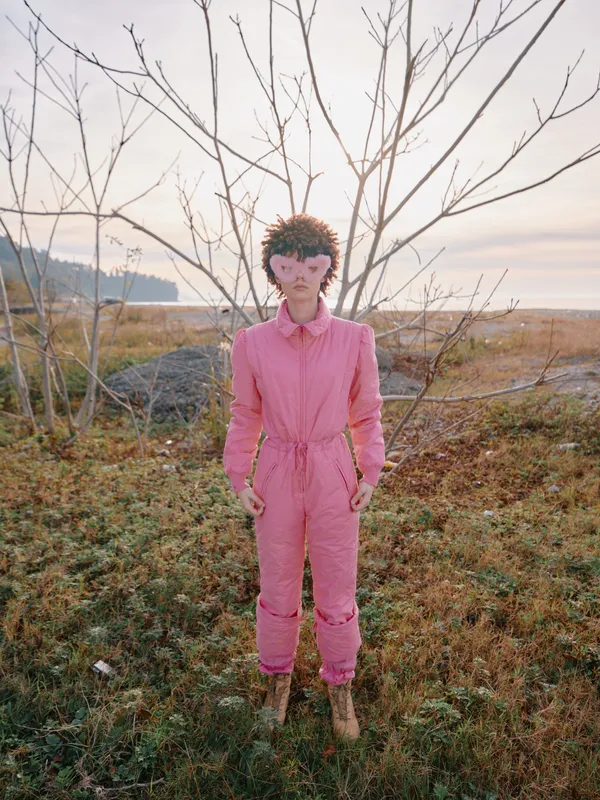 Pink jumpsuit woman stands in a field with bare trees, soft sunset light, an atmospheric outdoor portrait highlighting bold color, creative outfit and natural setting for fashion image use