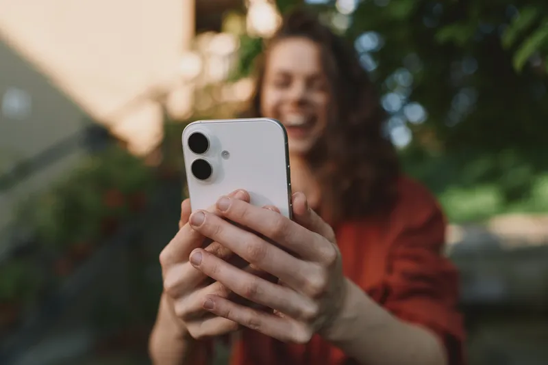 Young smiling woman in casual outfit takes a selfie outdoors with a smartphone, lifestyle shot capturing a candid moment, natural light and a blurred garden background