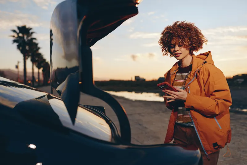 Woman with curly hair holding smartphone near car at sunset, outdoors lifestyle travel communication concept, warm golden light, casual jacket, soft evening mood, looking at screen