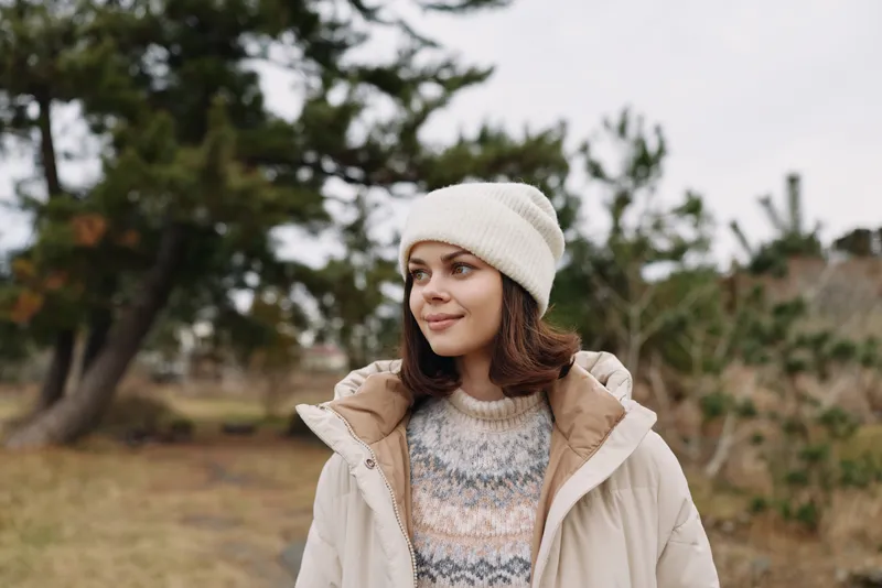 Portrait of a woman outdoors in a winter jacket and knit sweater, wearing a beanie, standing in a park with trees and muted autumn light.