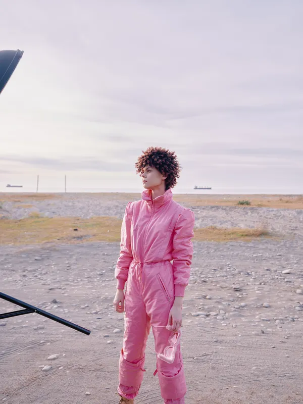 Pink jumpsuit worn by a model in an outdoor coastal setting, portrait style with wind in hair, soft light and expansive sky, showcasing contemporary fashion and street style with bold color and