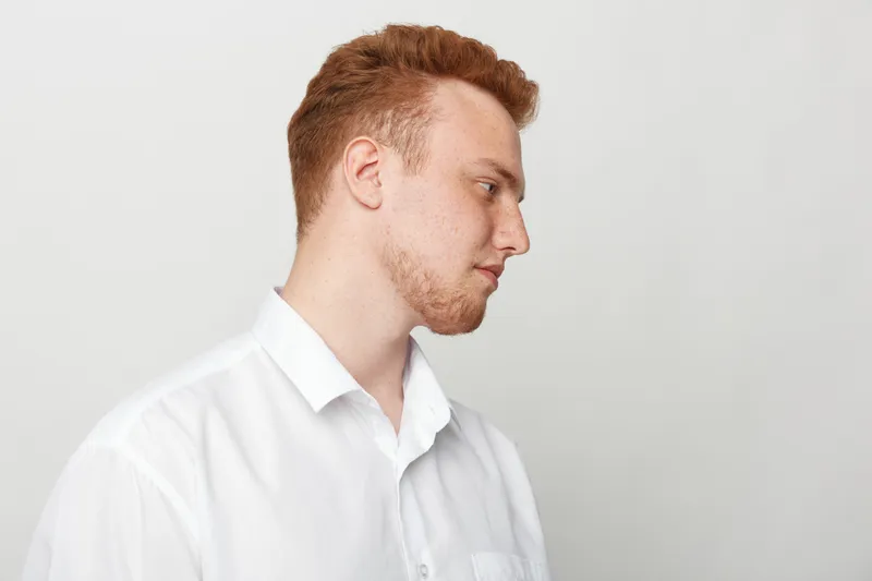 Profile of a young redhead man in a white shirt looking down with a neutral expression on a light grey background. Concept of contemplation, calmness, and casual style portrait.