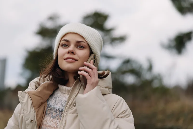 Woman in winter wear with a beige jacket and beanie, talking on a phone outdoors in natural light, portrait style, casual fashion mood amid calm scenery