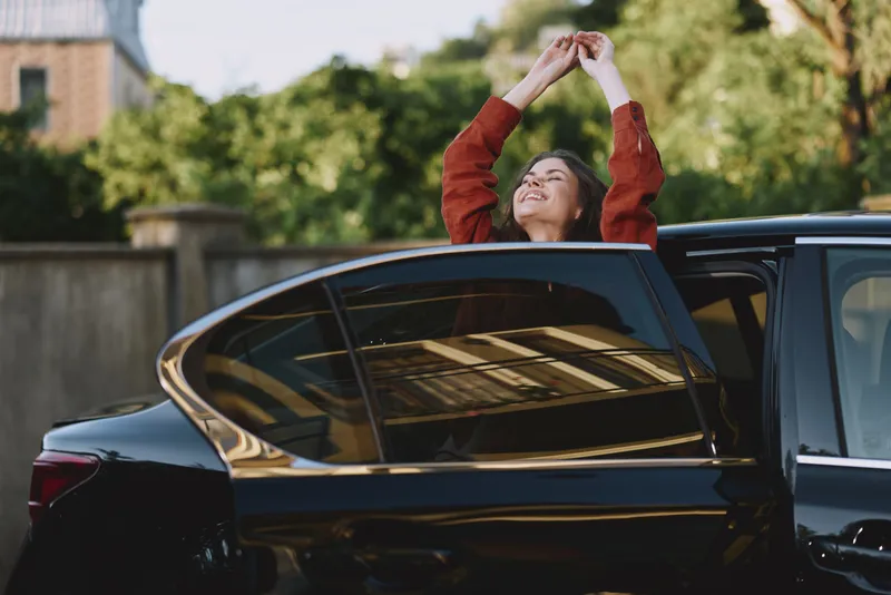 One woman leaning out of a sleek car window, smiling, wearing a red sweater and enjoying a candid moment during a casual city drive