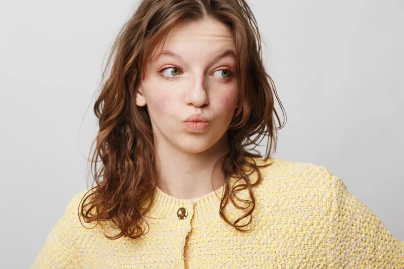 Young woman with curly hair wearing yellow sweater looks sideways with pursed lips. Studio portrait on light background. Concept of curiosity, doubt, and casual fashion.