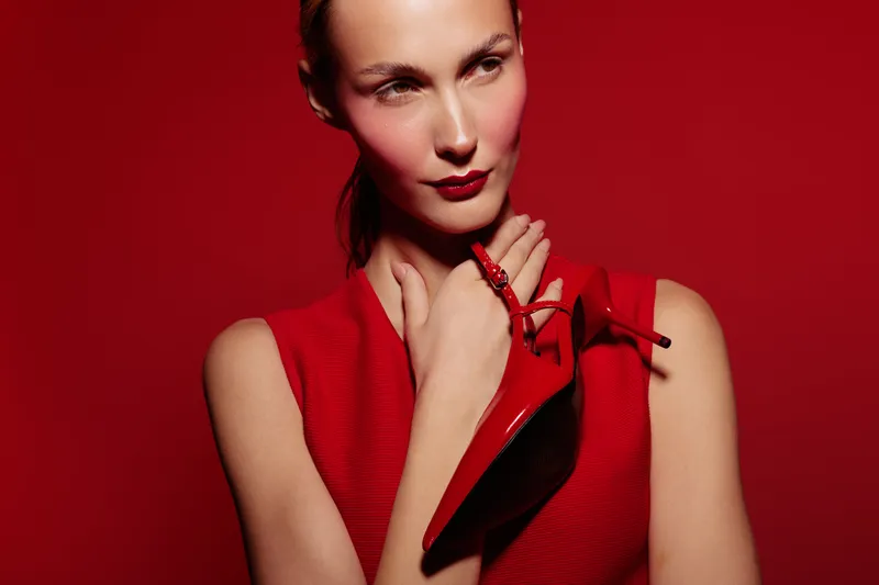 Young woman in red sleeveless top holds red high heel shoe near her neck against red background. Concept of fashion, style, elegance, and bold color contrast in studio lighting.