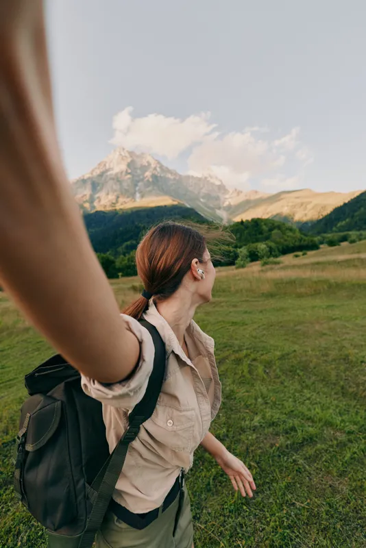 A young woman with a backpack walks across a green meadow toward distant mountains, pausing to admire rugged peaks and breathe in the fresh air.