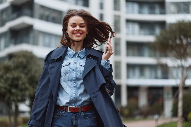Smiling woman wearing a denim jacket and shirt, standing outdoor with a modern city backdrop. Fashion portrait conveying confident casual energy and urban style.