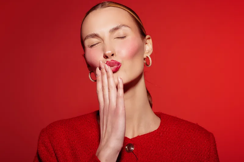 Young woman with closed eyes and red lips blowing a kiss on red background. She wears red clothing and gold hoop earrings. Concept of affection, beauty, and fashion portrait.