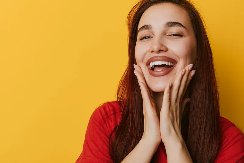 Young woman with long brown hair smiling and holding her face with both hands on yellow background. Portrait of happy female expressing joy and positive emotion with casual red shirt.