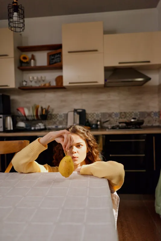 An introspective woman sits at a kitchen table, chin on hand, examining a lemon in front of her. A cozy home scene capturing casual, everyday life and domestic mood.