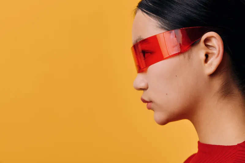 Portrait of young woman wearing red futuristic visor glasses with sleek hair in profile view against orange background. Stylish modern fashion and technology concept with minimalistic look.