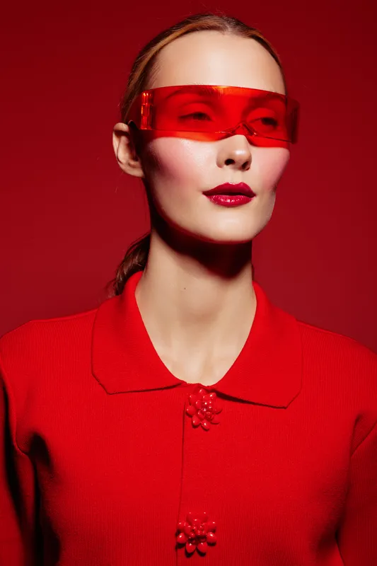 Stylish woman in red sweater and futuristic red sunglasses poses against red background. Concept of fashion, modern style, and bold color contrast in studio portrait.