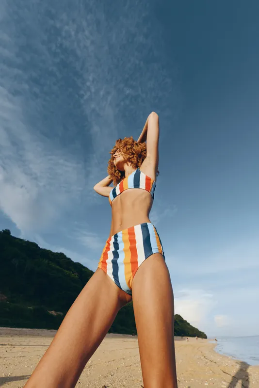 A young woman in a striped bikini stands on a sunny sandy beach, arms raised above her head, feeling the breeze as the bright sea stretches to the horizon and the coastline curves away.