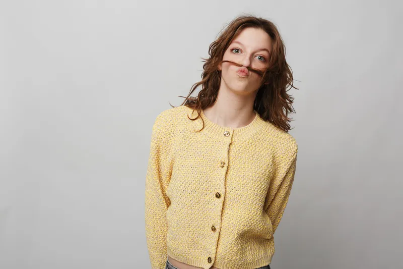 Young woman in yellow knitted sweater makes a playful mustache with her hair, standing against a plain light background. Concept of fun, humor, and casual lifestyle portrait.