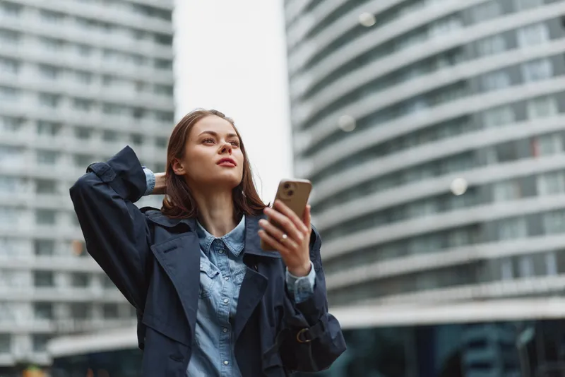 A young woman in a denim shirt and navy trench stands in a modern city plaza, looking up while holding a smartphone, conveying focus and contemplation amid glass towers and bustling streets.