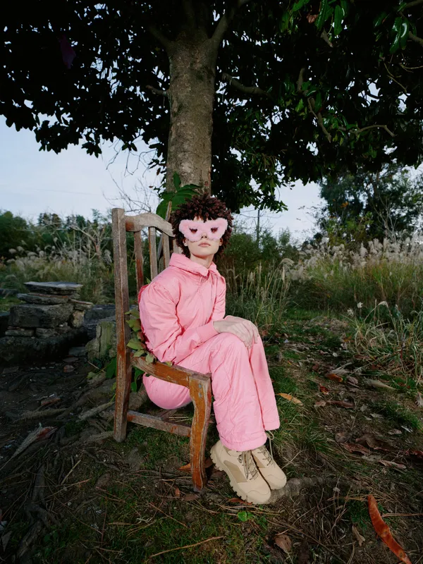 Outdoor portrait of a young girl in a pink jumpsuit seated on a wooden chair beneath a leafy tree, calm expression in a natural garden setting, soft light and quiet mood.