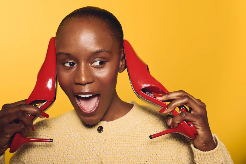 Happy dark skinned African woman holds red high heels near ears, smiling and looking away. Bright yellow background and casual sweater create lively and playful fashion concept.