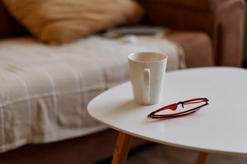 Close-up of a white mug on a round table with red glasses, a soft sofa in the background, creating a cozy home morning mood and casual lifestyle ambiance.