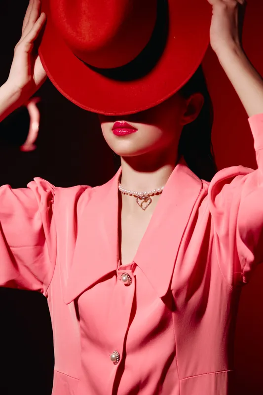 Fashionable woman in a stylish pink suit with red lipstick hides face behind a red hat. Creative beauty concept with dramatic lighting highlighting elegance and mystery.