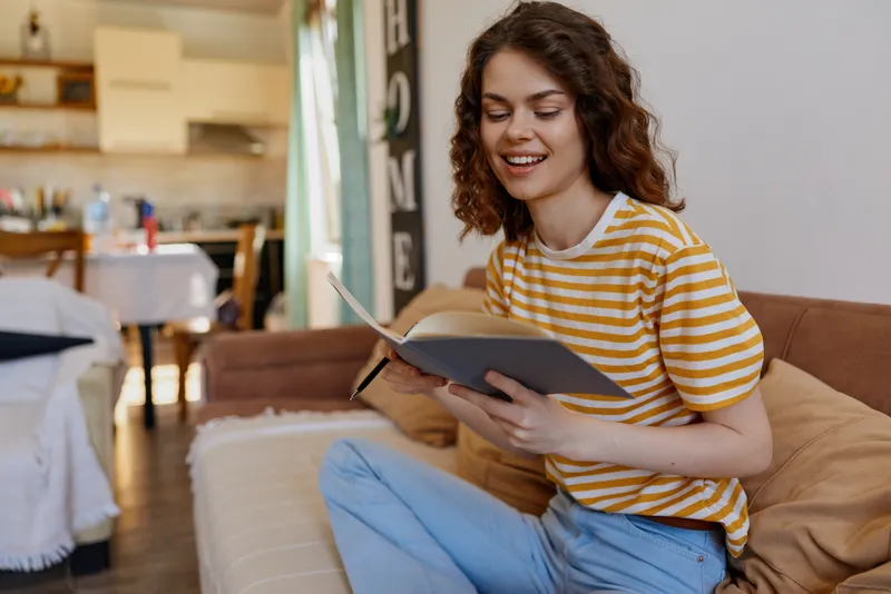 Smiling woman with curly hair wearing a striped shirt reads a notebook on a cozy couch at home, warm natural light, conveying lifestyle, comfort, and relaxation.
