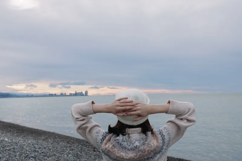 Person standing on a pebble beach facing the ocean with hands behind head, wearing a knitted sweater and hat, capturing a tranquil seaside moment.