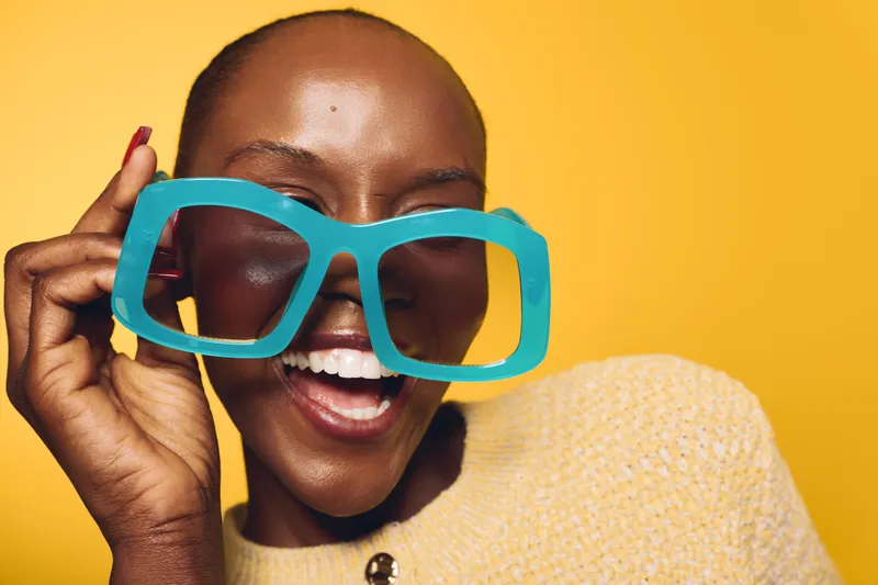 Happy African woman with dark skin laughing and holding oversized blue glasses near her face. Yellow background and cozy beige sweater create a cheerful and playful mood.